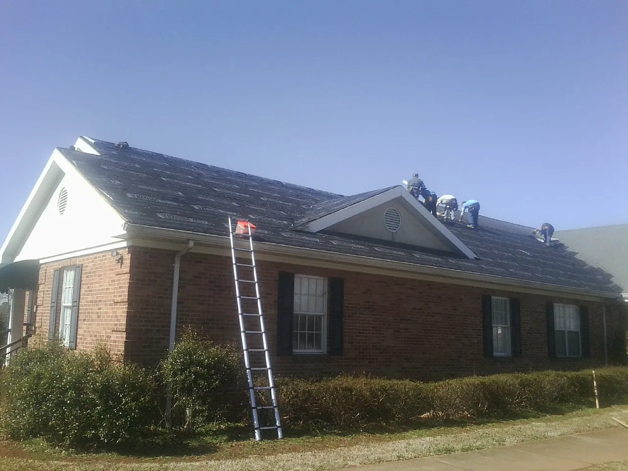 Crew tearing shingles off a brick ranch roof with a ladder set — Metal Roof Repair work by our crew in Roscoe