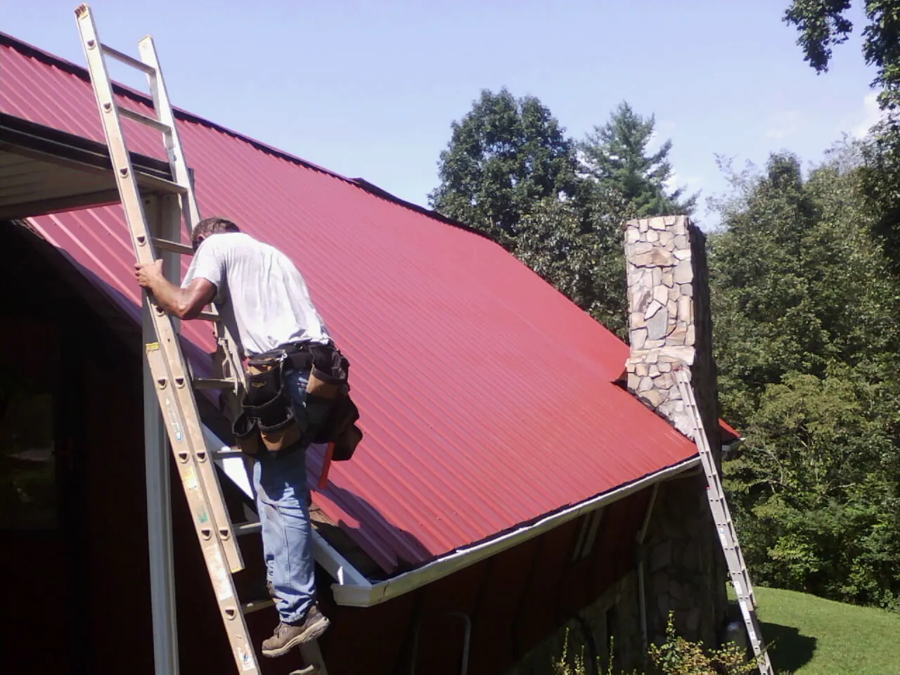 Roofer on a ladder beside a red metal roof and stone chimney — Soffit & Fascia Repair by Roofing Brothers in Roscoe